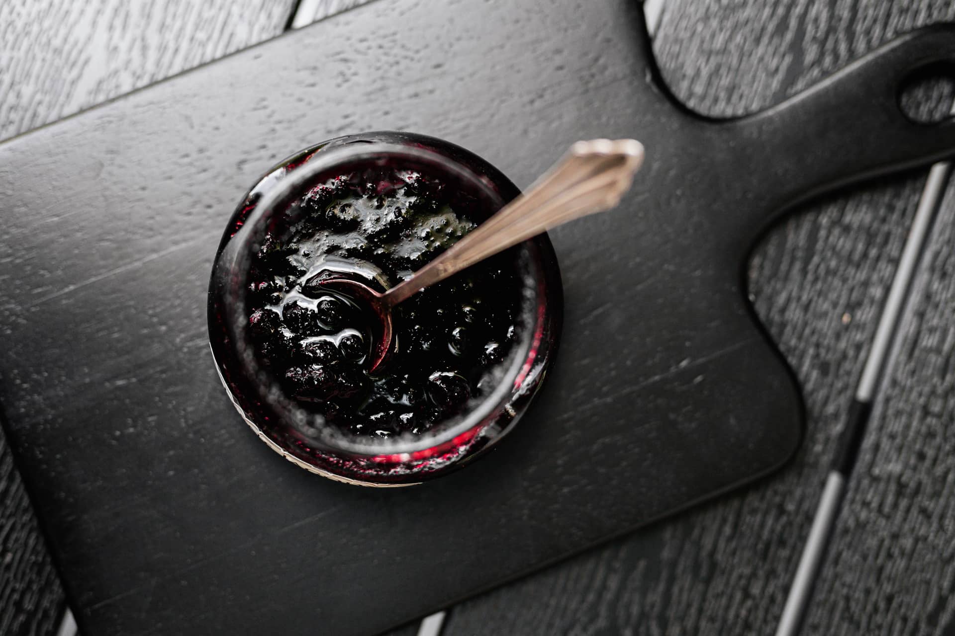 An open jar of dark fruit jam with a spoon on a black board